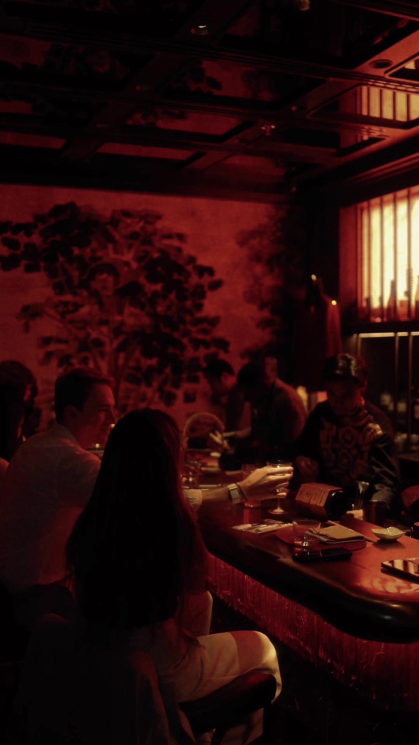 A night in Himitsu: the red-lit shelves behind the counter, a table laid with a bowl and chopsticks, and the room full.,ليلة في هيميتسو: الرفوف ذات الإضاءة الحمراء خلف الكاونتر، وطاولة عليها وعاء وعيدان، والغرفة ممتلئة.