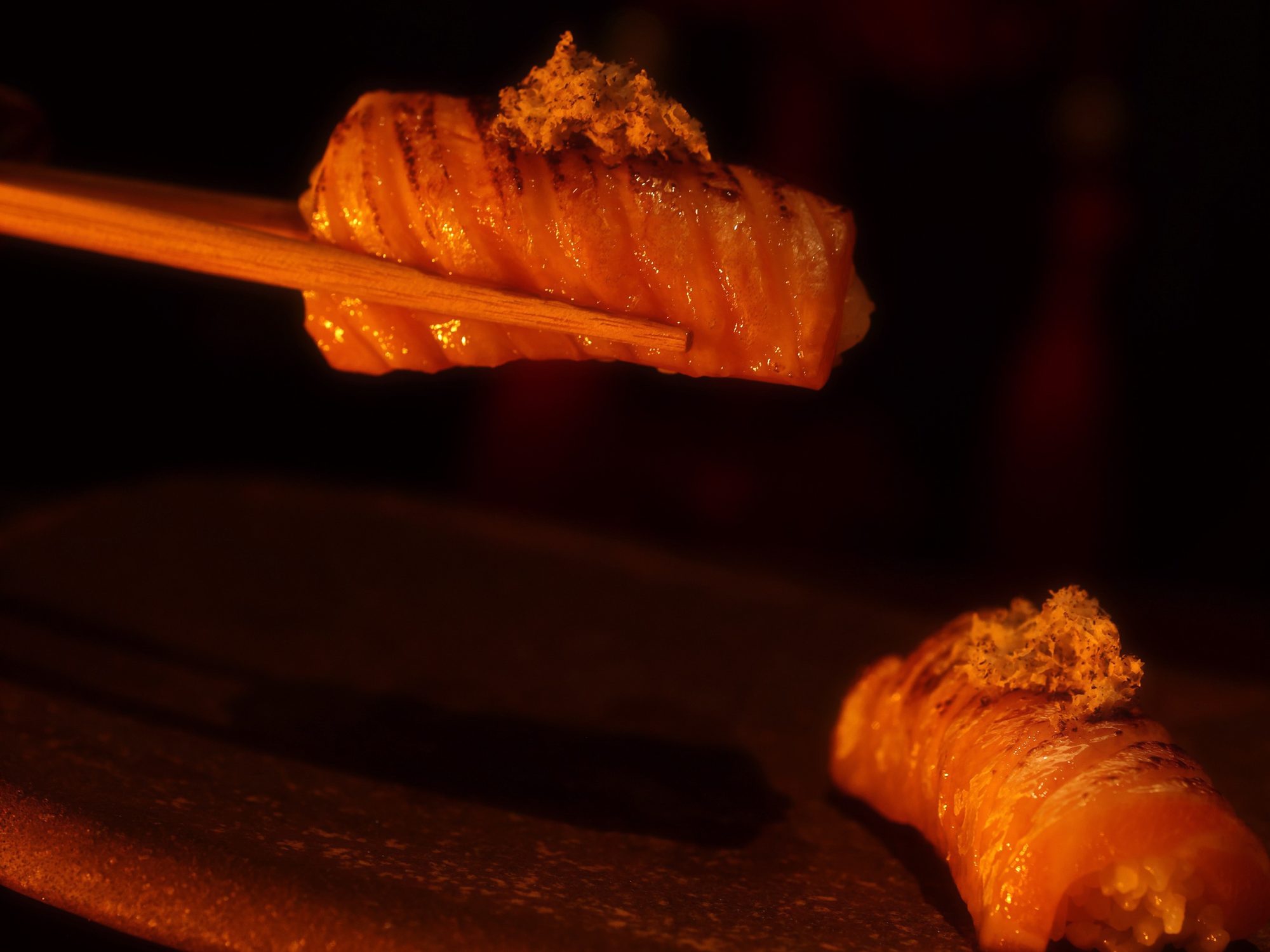 Two pieces of seared salmon nigiri under grated topping, one lifted on chopsticks in the red light of Himitsu.