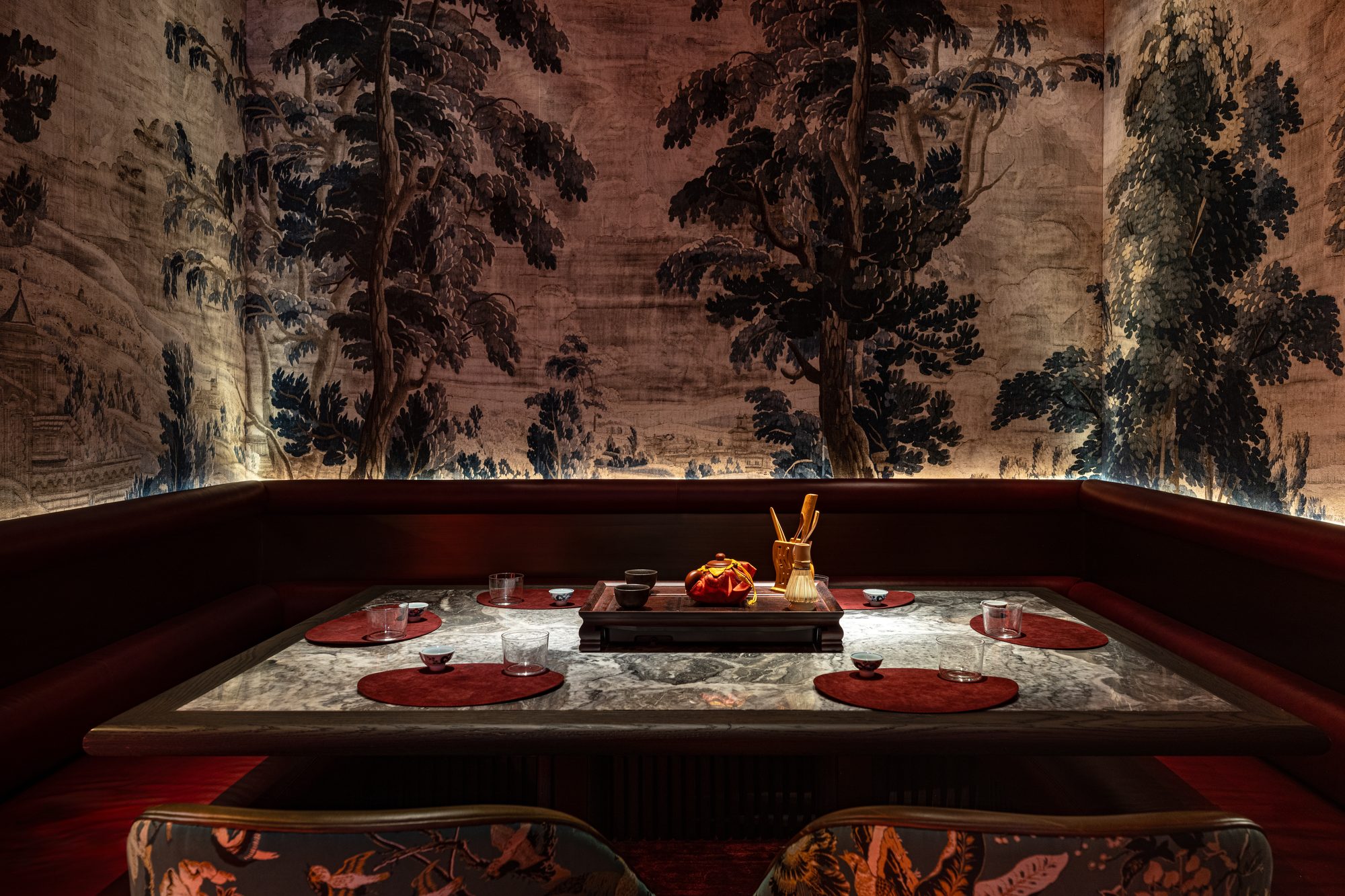 A corner booth in Himitsu: a red leather banquette under a hand-painted forest, a marble table laid for tea.