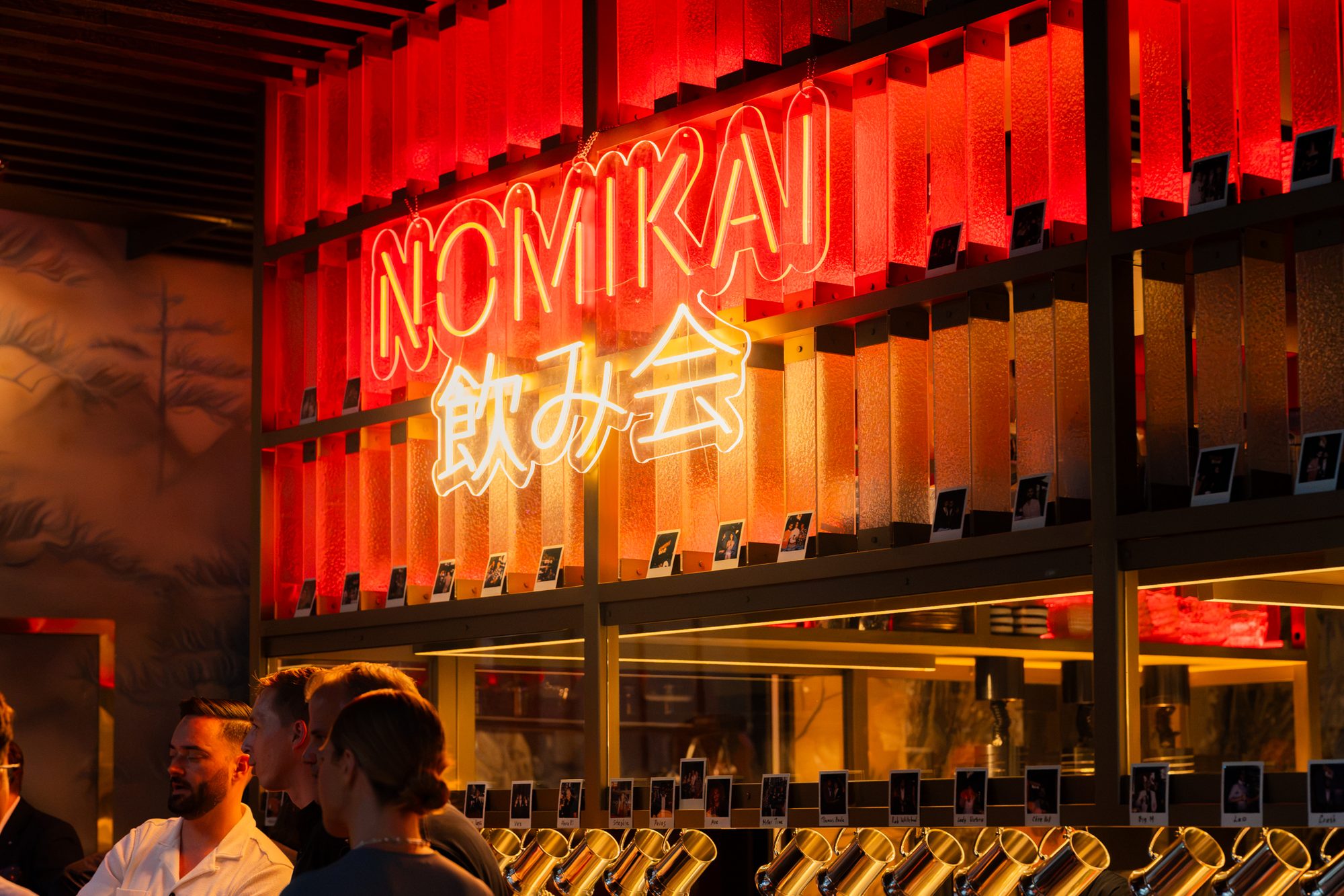 The Nomikai counter at Alba: a neon sign reading NOMIKAI 飲み会 above a wall of backlit panels hung with Polaroids, and a row of steins below.